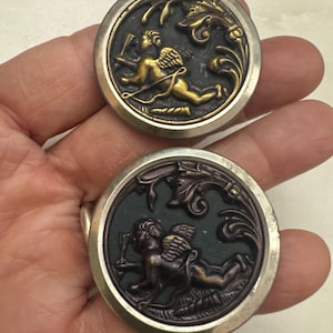 May include: Two antique buttons with a silver-colored rim. Each button features a cherub design in gold and bronze tones, surrounded by ornate floral details. The buttons are round and have a vintage aesthetic.