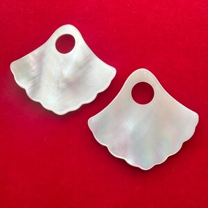 May include: Two iridescent white mother-of-pearl pendants with a fan-like shape and scalloped edges. Each pendant has a circular hole at the top. The pendants are displayed against a vibrant red background.