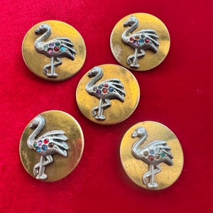 May include: Five vintage brass-toned buttons with a flamingo design. Each button features a silver-colored flamingo with colorful jewel accents on a gold-colored circular base. The buttons are arranged on a red background.