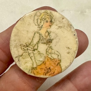 May include: A round, off-white vintage button featuring a hand-drawn illustration of a woman in a period dress. The woman's face is peach-colored, and her dress is a combination of white and orange. The button has a slightly aged appearance.