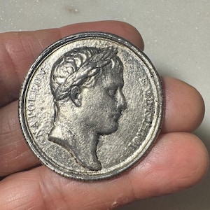 May include: A silver-colored coin featuring a profile of a person wearing a laurel wreath. The coin has the word "NAPOLEON" inscribed along the edge. The coin is held in a hand.