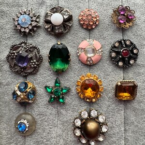 May include: A collection of decorative buttons in various shapes and colors. The buttons feature gemstones, rhinestones, and metal accents. Designs include floral, star, and geometric patterns. Colors include blue, pink, green, and amber.