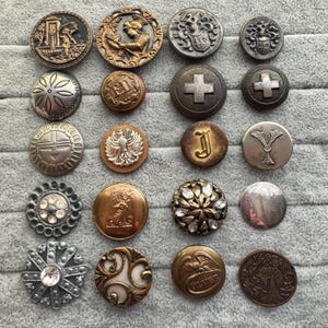 May include: A collection of antique buttons in various shapes, sizes, and designs. The buttons are made of metal and feature intricate details, including heraldic crests, floral patterns, and geometric shapes. Some buttons have a tarnished appearance, adding to their vintage charm.