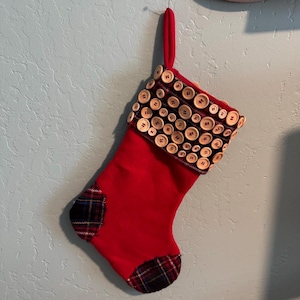 Può includere: Una calza di Natale rossa con tallone e punta a quadri. Il polsino è decorato con numerosi bottoni di legno. Un anello rosso è attaccato per appenderla. La calza è appesa contro un muro azzurro.