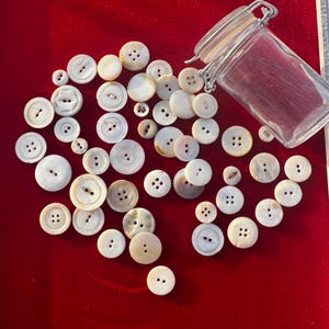 May include: A collection of various sized, off-white buttons with two or four holes, scattered on a red surface. A clear glass jar with a metal clasp is in the upper right corner. The buttons vary in size and shape.