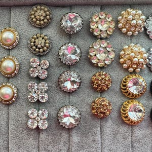 May include: A collection of decorative buttons in various designs. Some buttons feature clear rhinestones, pearls, and gold or silver-toned metal. Others have black or gold-colored beads. The buttons are arranged on a gray surface.