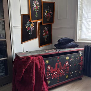 May include: A rectangular wooden storage chest with a black finish, decorated with red bird and floral motifs, featuring a hinged lid. A deep red fringed throw blanket hangs over the side, and four framed floral art pieces hang on the wall above.