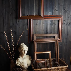 May include: Several wooden picture frames of different sizes, some mounted on a dark wood panelled wall. A classical bust, a wicker basket, and pine cones are also visible, creating a vintage aesthetic.