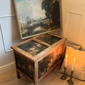 Peut inclure: Un coffre en bois avec une peinture de paysage sur le couvercle et le devant. Au-dessus, une peinture de paysage encadr&eacute;e est accroch&eacute;e au mur. Deux bougies allum&eacute;es dans des bougeoirs en laiton sont plac&eacute;es pr&egrave;s d'un coussin beige.