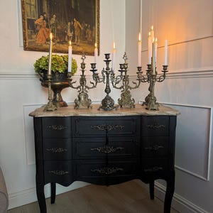 Puede incluir: Una cómoda antigua negra con tapa de mármol exhibe varios candelabros de latón ornamentados con velas encendidas. Una pintura enmarcada cuelga encima. La escena evoca una estética clásica y elegante.