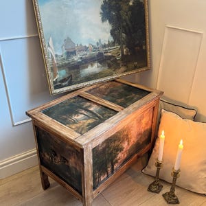 Peut inclure: Un coffre en bois avec une peinture de paysage sur le dessus et les c&ocirc;t&eacute;s. Le coffre a une finition marron fonc&eacute; et des poign&eacute;es en m&eacute;tal orn&eacute;. Une peinture de paysage encadr&eacute;e est accroch&eacute;e au-dessus. Deux bougies allum&eacute;es dans des bougeoirs en laiton sont &agrave; proximit&eacute;.