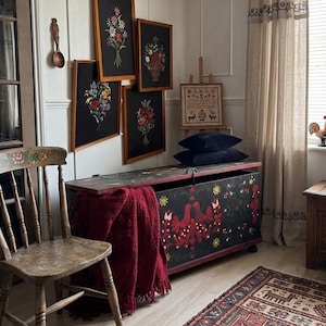 May include: A room featuring a wooden chair, a black storage chest with red floral folk art, a dark red throw blanket, four framed floral embroideries on black fabric, a small wooden easel with a sampler, and a patterned area rug on the floor.
