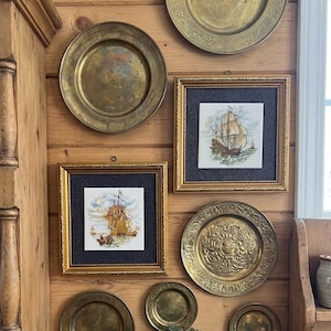 May include: A wall display featuring several round brass plates of varying sizes, two square framed prints depicting sailing ships, and a small shelf holding a dark book titled The Roux Brothers on PATISSERIE and a sprig of green pine needles.
