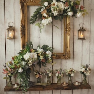 May include: A large ornate gold picture frame adorned with white and cream floral arrangements and green foliage. Below, a wooden shelf holds several small glass jars filled with assorted flowers, greenery, and two metal lanterns.