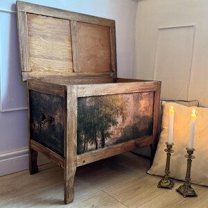 Peut inclure: Un coffre en bois antique avec un couvercle &agrave; charni&egrave;re, orn&eacute; d'une sc&egrave;ne de paysage peinte sur le devant. Le coffre a une finition vieillie, avec le grain du bois visible et une peinture sombre et d&eacute;taill&eacute;e. Deux bougeoirs en laiton avec des bougies allum&eacute;es sont &agrave; c&ocirc;t&eacute; du coffre.