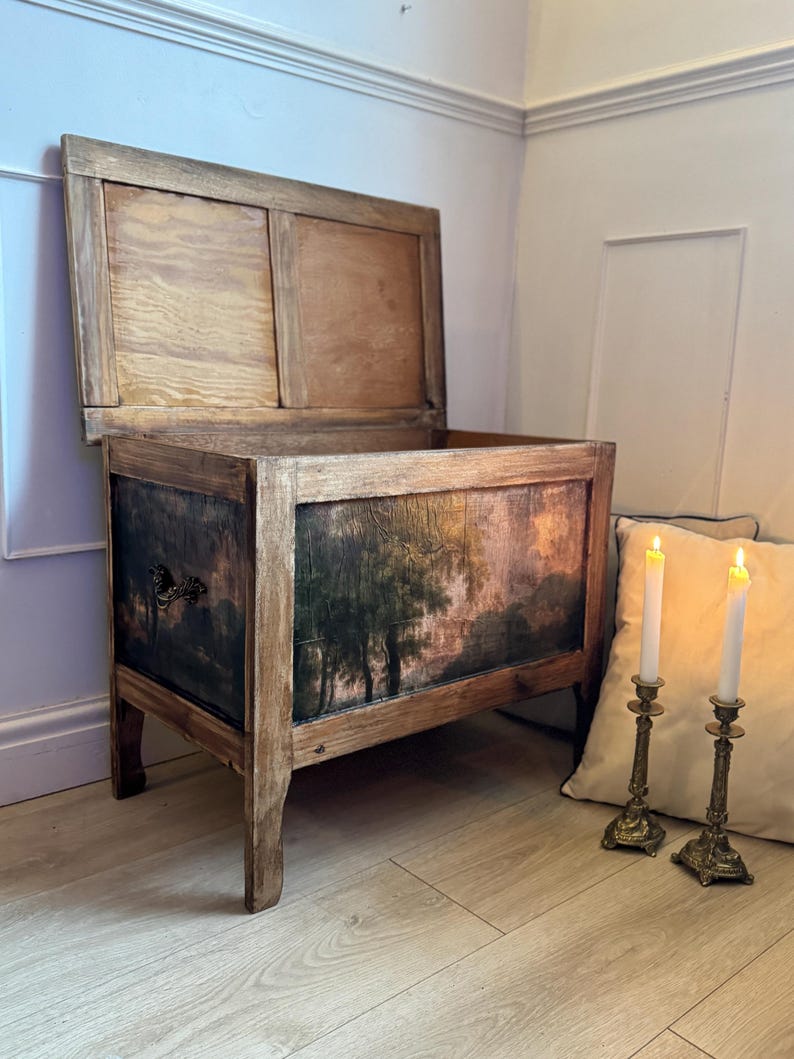 Peut inclure: Un coffre en bois antique avec un couvercle &agrave; charni&egrave;re, orn&eacute; d'une sc&egrave;ne de paysage peinte sur le devant. Le coffre a une finition vieillie, avec le grain du bois visible et une peinture sombre et d&eacute;taill&eacute;e. Deux bougeoirs en laiton avec des bougies allum&eacute;es sont &agrave; c&ocirc;t&eacute; du coffre.