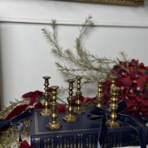 Peut inclure: Ensemble de chandeliers anciens en laiton disposés sur un livre bleu foncé. Des rubans de velours rouge et bleu marine et de la verdure entourent les bougeoirs. Un cadre doré est visible en arrière-plan.