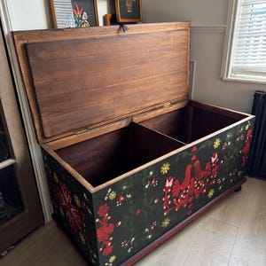 May include: A large rectangular wooden storage chest with an open lid, featuring a black exterior painted with red and yellow floral patterns and bird motifs. The interior is divided into two sections with natural wood grain panels and hinges.