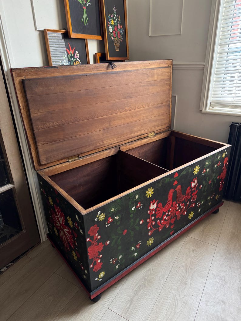 May include: A large rectangular wooden storage chest with an open lid, featuring a black exterior painted with red and yellow floral patterns and bird motifs. The interior is divided into two sections with natural wood grain panels and hinges.