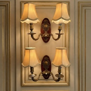 May include: Two double-arm wall sconces featuring brass-toned metal fixtures with ornate detailing, mounted on oval dark wood backplates. Each arm supports a cream-colored fabric lampshade with a scalloped bottom edge, emitting a warm light.