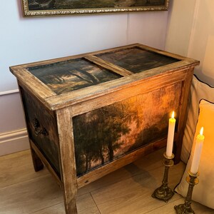 Peut inclure: Un coffre en bois avec une peinture de paysage sur les panneaux avant et sup&eacute;rieurs. Le coffre pr&eacute;sente une finition vieillie, avec un grain de bois visible et une peinture sombre et d&eacute;taill&eacute;e d'arbres. Deux bougies allum&eacute;es dans des bougeoirs orn&eacute;s sont &agrave; proximit&eacute;.