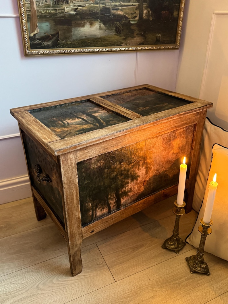 Peut inclure: Un coffre en bois avec une peinture de paysage sur les panneaux avant et sup&eacute;rieurs. Le coffre pr&eacute;sente une finition vieillie, avec un grain de bois visible et une peinture sombre et d&eacute;taill&eacute;e d'arbres. Deux bougies allum&eacute;es dans des bougeoirs orn&eacute;s sont &agrave; proximit&eacute;.