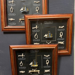 Peut inclure: Trois œuvres d'art nautiques encadrées. Chaque cadre présente un fond sombre avec divers nœuds de corde et un petit modèle de voilier. Les cadres sont en bois avec une finition marron foncé. Les œuvres sont exposées sur une surface gris foncé.