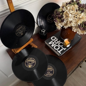 Pode incluir: Vários discos de vinil pretos com rótulos dourados são exibidos em uma mesa de madeira. Um disco está apoiado em um suporte de madeira. Um vaso de flores secas, uma vela e um livro também estão na mesa.