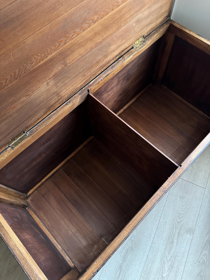 May include: A rectangular wooden storage chest with an open hinged lid, featuring a natural brown wood grain finish and a central vertical divider that separates the interior into two distinct storage compartments with visible metal hinges.