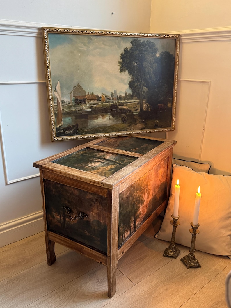 Peut inclure: Un coffre en bois ancien avec une peinture de paysage sur le devant et le dessus. Le coffre est rectangulaire avec quatre pieds. Une peinture de paysage encadr&eacute;e est accroch&eacute;e au-dessus. Deux bougies allum&eacute;es dans des bougeoirs en laiton sont &agrave; proximit&eacute;.