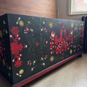 May include: A rectangular black storage chest featuring hand-painted folk art motifs in red, green, and yellow. The design includes symmetrical floral patterns and two facing birds. The chest rests on a red base with small, dark, rounded feet.