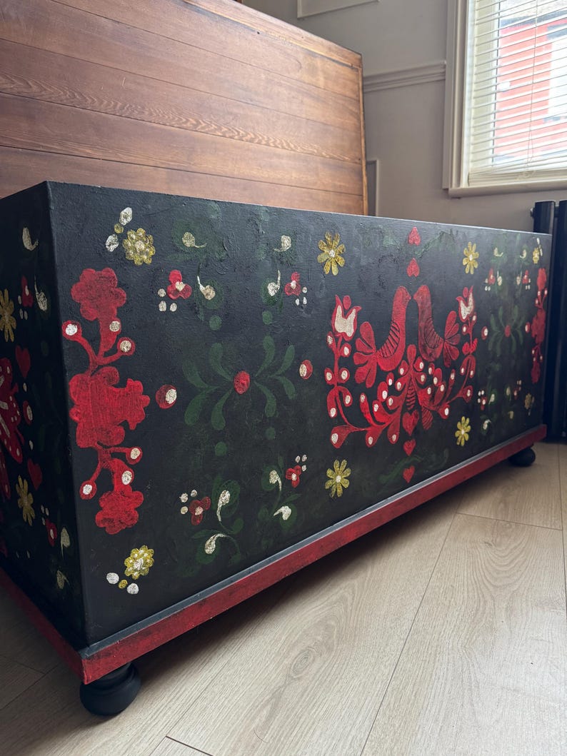 May include: A rectangular black storage chest featuring hand-painted folk art motifs in red, green, and yellow. The design includes symmetrical floral patterns and two facing birds. The chest rests on a red base with small, dark, rounded feet.