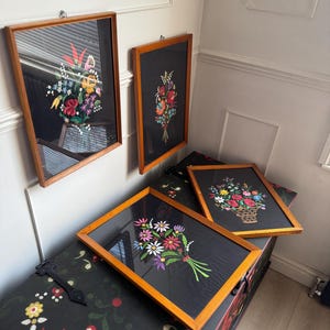 May include: Four rectangular framed wall art pieces featuring colorful floral embroidery on black fabric. Each piece is encased in a light brown wooden frame and displays a unique bouquet of flowers in shades of red, yellow, blue, and green.