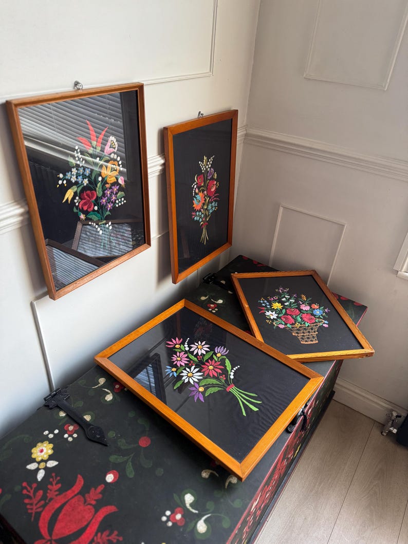 May include: Four rectangular framed wall art pieces featuring colorful floral embroidery on black fabric. Each piece is encased in a light brown wooden frame and displays a unique bouquet of flowers in shades of red, yellow, blue, and green.