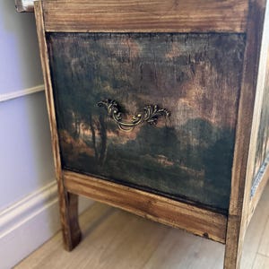 Peut inclure: Une commode en bois avec une peinture de paysage sur le panneau avant. Le coffre a une apparence sombre et vieillie avec une poign&eacute;e d&eacute;corative en laiton. Le cadre est en bois clair.