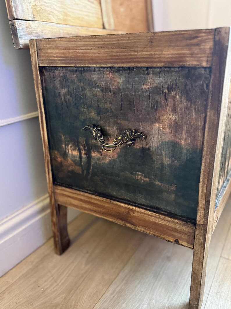 Peut inclure: Une commode en bois avec une peinture de paysage sur le panneau avant. Le coffre a une apparence sombre et vieillie avec une poign&eacute;e d&eacute;corative en laiton. Le cadre est en bois clair.