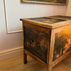 Peut inclure: Un coffre en bois vintage avec une peinture de paysage sur le devant et le dessus. Le coffre a un aspect sombre et vieilli avec une poign&eacute;e d&eacute;corative. Le cadre est en bois de couleur claire.