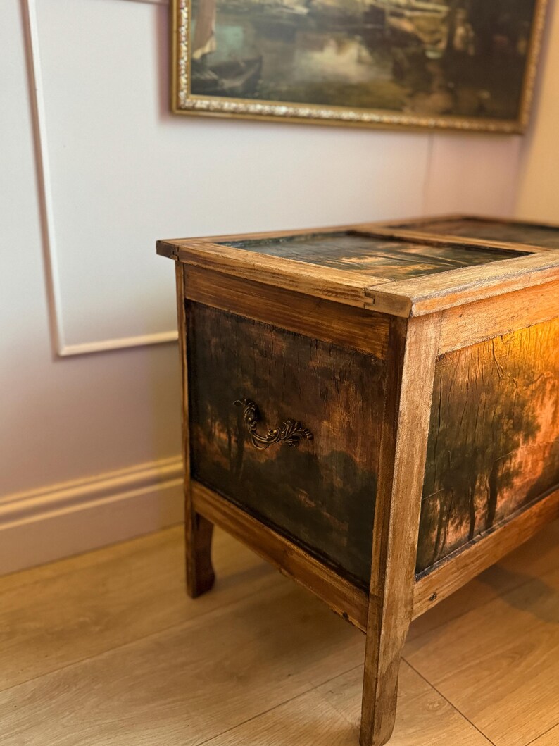Peut inclure: Un coffre en bois vintage avec une peinture de paysage sur le devant et le dessus. Le coffre a un aspect sombre et vieilli avec une poign&eacute;e d&eacute;corative. Le cadre est en bois de couleur claire.