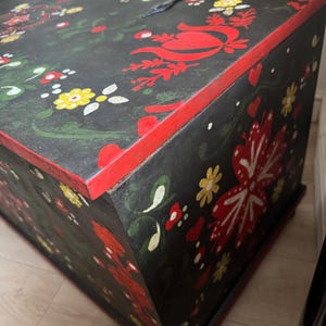 May include: A rectangular wooden chest with a black base color, featuring hand-painted floral motifs in red, yellow, and green. The edges are highlighted in red, and the lid is secured with black metal hinges. The surface displays intricate patterns of stylized flowers, leaves, and small heart shapes.