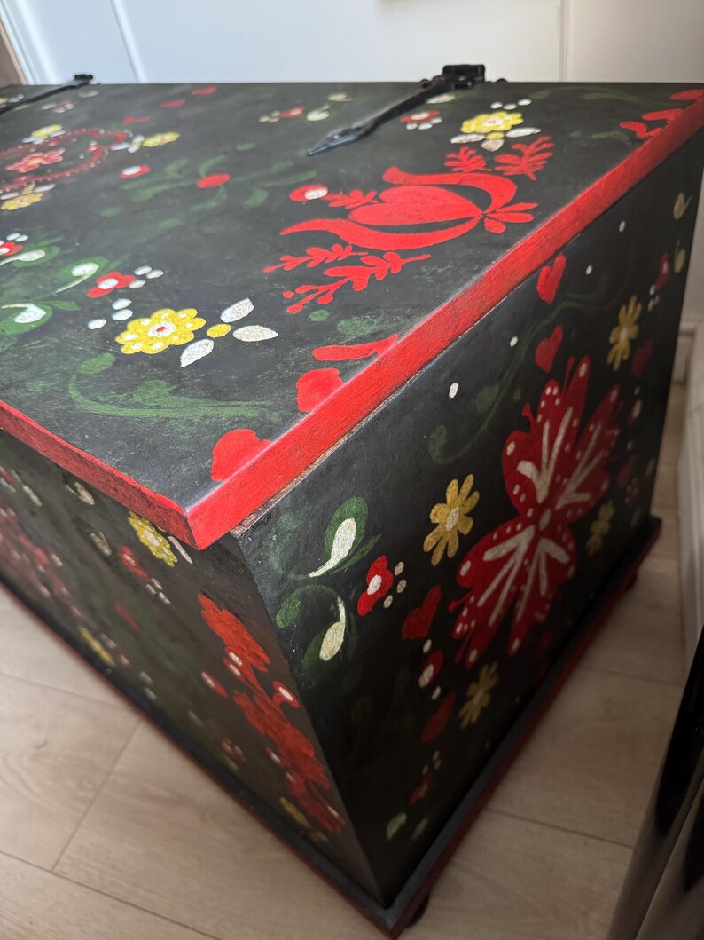 May include: A rectangular wooden chest with a black base color, featuring hand-painted floral motifs in red, yellow, and green. The edges are highlighted in red, and the lid is secured with black metal hinges. The surface displays intricate patterns of stylized flowers, leaves, and small heart shapes.