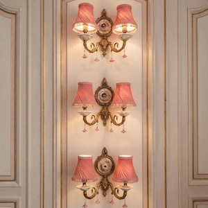 May include: Three matching wall sconces mounted vertically on a panelled wall. Each fixture features two pink pleated fabric shades with fringe, gold-toned metal scrollwork, a central circular porcelain medallion, and small dangling pink crystal accents.