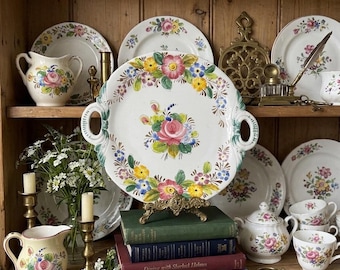 May include: A round ceramic serving platter with two handles, featuring a hand-painted floral motif with pink, yellow, and blue flowers on a white background. The platter is displayed on a stack of four books, surrounded by various floral-patterned dishes.