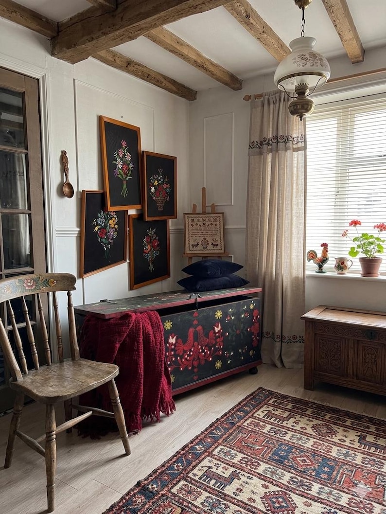 May include: A room featuring a wooden chair, a black storage chest with red floral folk art, a dark red throw blanket, four framed floral embroideries on black fabric, a small wooden easel with a sampler, and a patterned area rug on the floor.