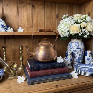 May include: A polished copper tea kettle with a wooden handle sits atop a stack of three hardcover books. The books are titled Dining with Sherlock Holmes and Roux Brothers on PATISSERIE. The kettle is surrounded by white flowers and brass items.