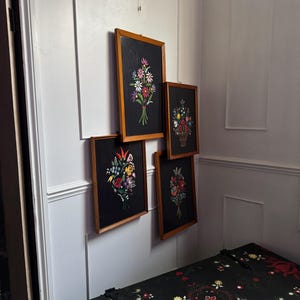 May include: Four rectangular wall art pieces in light wood frames, each featuring a black background with a colorful floral bouquet illustration. The flowers are depicted in shades of red, white, yellow, blue, and pink with green stems and leaves.