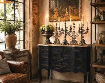 May include: A black wooden chest of drawers with three drawers featuring ornate gold-toned metal pulls and curved legs. The chest is topped with a light-colored stone surface holding four intricate metal candelabras with lit white taper candles.