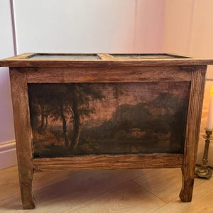 Peut inclure: Un coffre en bois avec une peinture de paysage sur le devant. Le coffre est de forme rectangulaire avec un couvercle &agrave; charni&egrave;re et quatre pieds. La peinture repr&eacute;sente des arbres, de l'eau et un b&acirc;timent, dans des tons de brun, noir et beige.