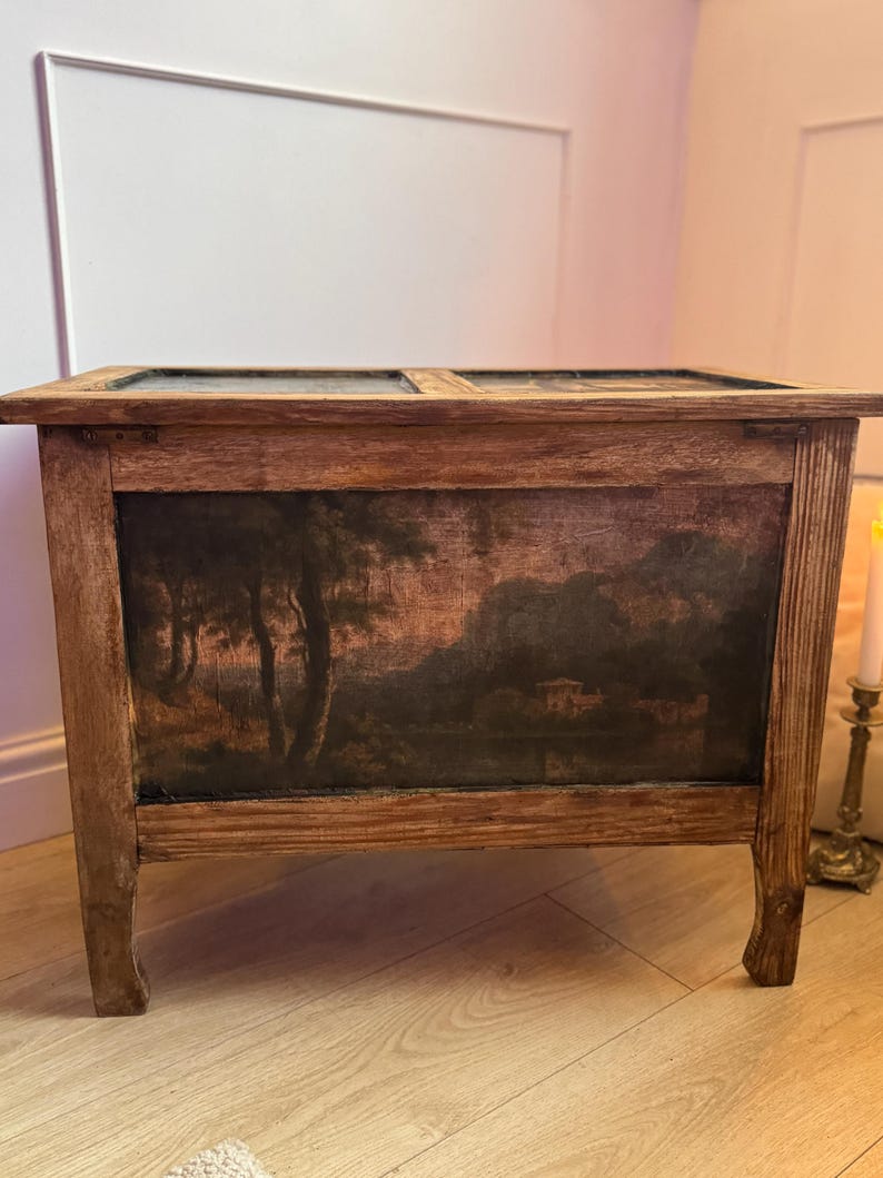 Peut inclure: Un coffre en bois avec une peinture de paysage sur le devant. Le coffre est de forme rectangulaire avec un couvercle &agrave; charni&egrave;re et quatre pieds. La peinture repr&eacute;sente des arbres, de l'eau et un b&acirc;timent, dans des tons de brun, noir et beige.