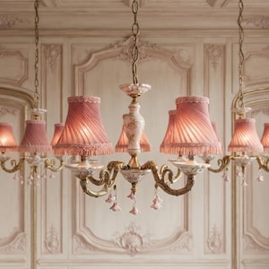 May include: A multi-armed chandelier featuring brass scrollwork arms, a central ceramic column with floral motifs, and pleated pink fabric lampshades with fringed edges. Small pink ceramic flower pendants hang from the base of each light fixture.