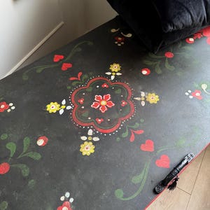 May include: A black wooden storage chest featuring a hand-painted floral motif in red, yellow, and green. The design includes stylized flowers, leaves, and heart shapes across the top surface, which also has a black metal handle attached to the side.